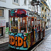 Straßenbahn mit Graffiti, die auf einer abschüssigen Straße mit Fußgängern und Wohnhäusern bergab fährt; Lissabon, Region Lisboa, Portugal
