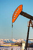 Close-up of pumpjack with the warm light at sunrise, snow-covered mountains and blue sky in the background, West of Airdrie; Alberta, Canada