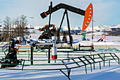 Brunnenkopf mit Pipeline und Pumpjack in einem schneebedeckten Feld mit schneebedeckten Bergen und blauem Himmel im Hintergrund, nördlich von Longview; Alberta, Kanada