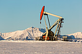 Pumpjack in a snow-covered field with snow-covered mountains and blue sky in the background; Longview, Alberta, Canada