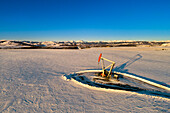 Luftaufnahme eines Pumpjacks in einem schneebedeckten Feld mit langen Schatten, warmem Licht des Sonnenaufgangs, sanften Hügeln, schneebedeckten Bergen und blauem Himmel im Hintergrund; Longview, Alberta, Kanada