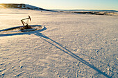 Aerial view of pumpjack in a snow-covered field with long shadows, warm light of sunrise, rolling hills, snow-covered mountains and blue sky in the background; Longview, Alberta, Canada