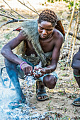 Junger Hadzabe-Jäger mit Pavianfellmantel rupft einen bei der morgendlichen Jagd erlegten Vogel und bereitet sich darauf vor, ihn über einem offenen Feuer in der Nähe des Eyasi-Sees zu braten; Tansania
