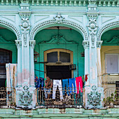 Ein Gebäude mit verschnörkelter und dekorativer Fassade und einer Wäscheleine, die über einem Balkon hängt, auf dem ein Mann sitzt und hinausschaut; Havanna, Kuba