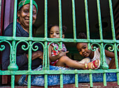 A mother with two young daughters sits looking out bars from their home; Havana, Cuba