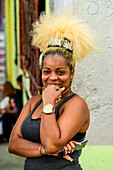 A Cuban woman with a unique blond hairstyle stands holding her smart phone and posing for the camera; Havana, Cuba