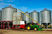 Silos im Hintergrund mit einem Traktor und einer Schnecke zum Beladen eines Getreidetrockners im Vordergrund eines Bauernhofs; Legal, Alberta, Kanada