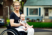 Eine querschnittsgelähmte Mutter, die ihr Baby auf dem Schoß trägt, während sie an einem warmen Sommernachmittag im Freien einen Rollstuhl benutzt: Edmonton, Alberta, Kanada