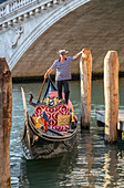 Gondolier stands on the back of a gondola at the waterfront waiting for tourists; Venice, Italy