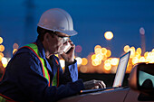 Mann, der an einem Laptop und einem Smartphone arbeitet, mit einer Ölraffinerie im Hintergrund; Alberta, Kanada