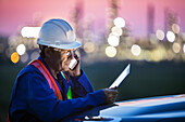 Man working on a tablet and smart phone with an oil refinery in the background; Alberta, Canada