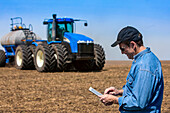 Landwirt benutzt ein Tablet, während er auf einem landwirtschaftlichen Feld steht und einen Traktor und Maschinen beim Säen des Feldes beobachtet; Alberta, Kanada