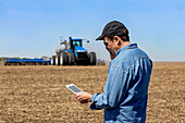 Landwirt, der ein Smartphone und ein Tablet benutzt, während er auf einem landwirtschaftlichen Feld steht und den Traktor und die Maschinen beim Säen des Feldes beobachtet; Alberta, Kanada