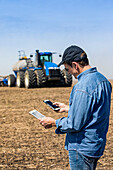 Ein Landwirt benutzt ein Smartphone und ein Tablet, während er auf einem landwirtschaftlichen Feld steht und den Traktor und die Geräte beim Säen des Feldes beobachtet; Alberta, Kanada