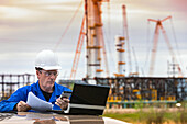 Man works on laptop and smart phone while standing at a vehicle with an oil rig in the background; Alberta, Canada
