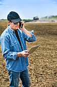 Landwirt, der ein Smartphone und ein Tablet benutzt, während er auf einem landwirtschaftlichen Feld steht und den Traktor und die Maschinen beim Säen des Feldes beobachtet; Alberta, Kanada