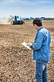 Ein Landwirt benutzt ein Tablet, während er auf einem landwirtschaftlichen Feld steht und einen Traktor und Geräte beobachtet, die das Feld säen; Alberta, Kanada