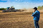 Landwirt, der ein Smartphone und ein Tablet benutzt, während er auf einem Feld steht und den Traktor und die Geräte beobachtet, die das Feld säen; Alberta, Kanada