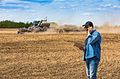 Ein Landwirt benutzt ein Smartphone und ein Tablet, während er auf einem landwirtschaftlichen Feld steht und den Traktor und die Geräte bei der Aussaat beobachtet; Alberta, Kanada