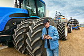 Landwirt, der ein Smartphone benutzt, während er auf einem Bauernhof neben den Geräten steht; Alberta, Kanada