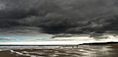 Dunkle Gewitterwolken über dem Atlantischen Ozean mit zwei Menschen und ihrem Hund, die am nassen Sandstrand im Vordergrund spazieren gehen; South Shields, Tyne and Wear, England