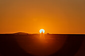 Man stands backlit by the bright, glowing sun as it sinks behind the horizon at sunset in Aluvlei, Namib-Naukluft National Park; Namibia