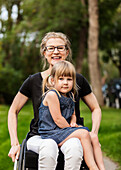 A paraplegic mom holding her little girl in her lap while sitting in her wheelchair in her front yard on a warm summer afternoon: Edmonton, Alberta, Canada.