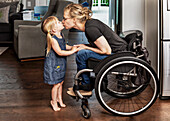 Ein junges Mädchen stellt sich auf die Zehenspitzen, um ihrer querschnittsgelähmten Mutter, die im Rollstuhl sitzt, einen Kuss zu geben: Edmonton, Alberta, Kanada.