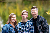 A young man with Down Syndrome posing for a family portrait with his father and mother while enjoying each other's company in a city park on a warm fall evening: Edmonton, Alberta, Canada