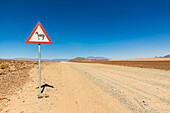Schild "Achtung Wildtiere" an einer langen Trockenstraße, Namib-Wüste, Namib-Naukluft-Nationalpark; Namibia