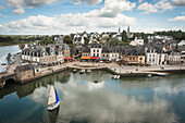 A small sailing boat with a blue and white sail approaches the harbour of Saint Gustan, an old port town with traditional buildings; Saint Gustan, Brittany, France