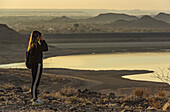 Female tourist looking out at Hardap Dam with binoculars at sunrise; Hardap Region, Namibia