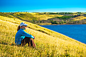 Frau sitzt auf einem grasbewachsenen Hügel mit Blick auf einen See und sanfte Hügel im Hintergrund mit blauem Himmel, Waterton Lakes National Park; Waterton, Alberta, Kanada