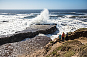 Drei Personen stehen auf einer Klippe und blicken auf die Wellen, die gegen die Felsen unter ihnen schlagen, Wilder Ranch State Park; Kalifornien, Vereinigte Staaten von Amerika