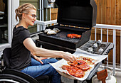 A paraplegic woman cooking steaks and baked potatoes on an outdoor barbecue while sitting in her wheelchair: Edmonton, Alberta, Canada
