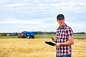Ein Landwirt nutzt sein Tablet, um die Weizenernte zu verwalten, während im Hintergrund ein Getreidewagen arbeitet; Alcomdale, Alberta, Kanada