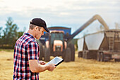 Ein Landwirt nutzt sein Tablet, um die Weizenernte zu verwalten, während ein Mähdrescher eine volle Ladung Getreide auf einen Getreidewagen ablädt: Alcomdale, Alberta, Kanada