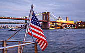 Eine amerikanische Flagge weht an einem Geländer an der Uferpromenade mit Blick auf die Brooklyn Bridge, Manhattan; New York City, New York, Vereinigte Staaten von Amerika