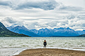 Woman stands on a sandy beach looking out at Carcross Lake and the vast Yukon Ranges; Carcross, Yukon, Canada