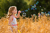 Junges Mädchen steht in einem goldenen Weizenfeld und bläst Seifenblasen; Alberta, Kanada
