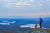 Lone woman hiker with knit hat and dog looking at each other on a cliff edge overlooking lakes in the distance on a cloudy evening in winter, Burren National Park; County Clare, Ireland