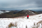 Einsame Wanderin in roter Jacke beim Aufstieg auf einen schneebedeckten Berg an einem bewölkten Wintertag, Galty Mountains; Grafschaft Tipperary, Irland