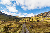 Alte irische Schotterstraße, die zu steilen Klippen und Bergen führt, im Sonnenlicht an einem sonnigen Tag mit Wolken am Himmel; Eagles Rock, County Leitrim, Irland