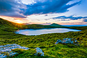 Sun setting over Lough Muskery in the Galty Mountains in summer with large boulders in the foreground, Galty Mountains; County Limerick, Ireland
