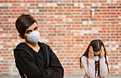 Siblings wearing protective masks to protect against COVID-19 during the Coronavirus World Pandemic, the boy with arms crossed and the girl with hands holding her head in fear; Toronto, Ontario, Canada