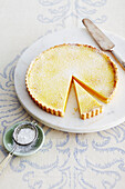 Lemon Tart on Marble Stand with Pie Server and Bowl of Icing Sugar on Table in Studio