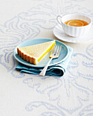 Scheibe Zitronenkuchen und Gabel auf blauem Teller mit Tasse und Untertasse mit Kräutertee auf Tischdecke im Studio