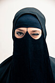 Close-up portrait of young woman wearing black, muslim hijab and muslim dress, winking and looking at camera, eyes showing eye makeup, studio shot on white background