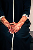 Woman sitting indoors with close-up of arms and hands painted with henna in arabic style, wearing a typical black, arabic, muslim dress, studio shot