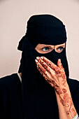 Nahaufnahme einer Frau, die einen schwarzen muslimischen Hidschab und ein muslimisches Kleid trägt und zur Seite schaut, wobei sie den Mund mit der Hand bedeckt und Arme und Hände zeigt, die mit Henna im arabischen Stil bemalt sind, Studioaufnahme vor weißem Hintergrund